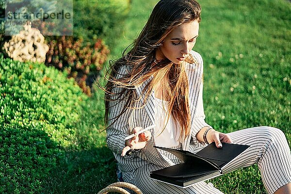 Junge schöne Frau entspannt sich im Stadtpark und macht sich Notizen in ihrem Notizbuch auf dem grünen Rasen. Ruhe und Freizeit Konzept