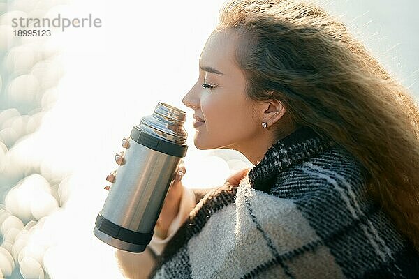 Junge hübsche Frau hält eine Thermoskanne und trinkt Tee in Decke eingewickelt über Unschärfe Fluss Hintergrund. Entspannen Konzept  glücklich Herbst