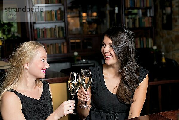 Zwei schöne elegante Frauen Freunde in stilvollen einfachen schwarzen Cocktailkleider Toast einander mit Gläsern von gekühlten Champagner  wie sie auf eine Nacht zusammen feiern