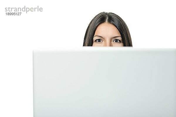 Junge Frau  die mit intelligenten Augen in die Kamera schaut  während sie sich hinter dem Monitor eines Tischcomputers versteckt  mit Kopierraum auf weißem Grund