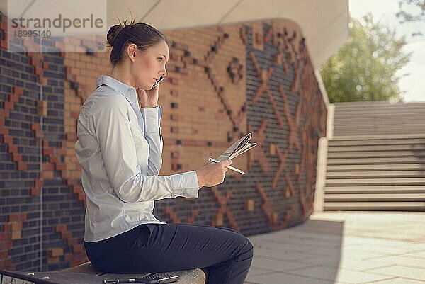 Frau  die in einem kommerziellen Foyer auf einer an der Wand befestigten Bank vor gemustertem Mauerwerk sitzt und mit ihrem Mobiltelefon spricht und ihre Notizen mit einem Tablet daneben bespricht  Konzept der Mobilität