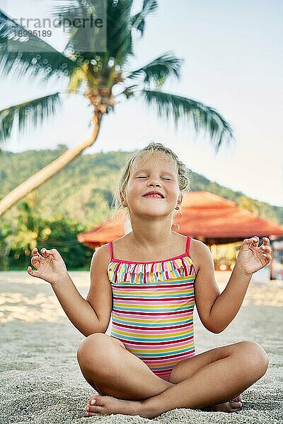 Hübsches kleines Mädchen meditiert in Lotuspose am Sandstrand im Sommerurlaub. Glückliche Kindheit Konzept