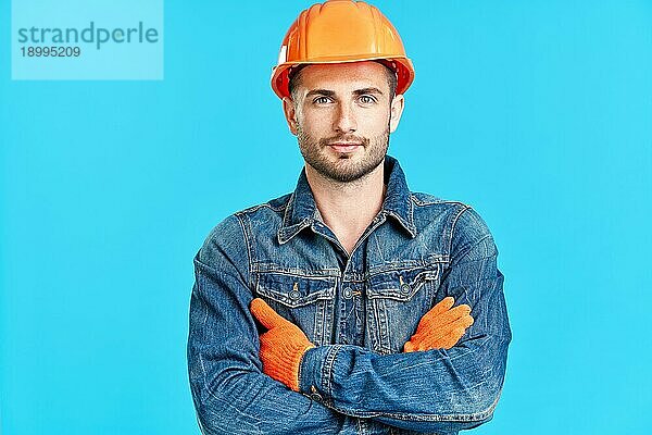 Porträt eines selbstbewussten  gut aussehenden männlichen Bauarbeiters mit Sicherheitshelm und verschränkten Armen  der auf einem blaün Hintergrund steht und zur Kamera schaut