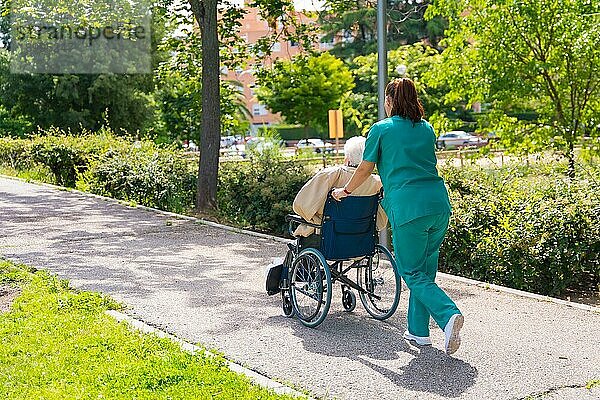 Eine ältere Frau mit der Krankenschwester bei einem Spaziergang im Garten eines Pflegeheims im Rollstuhl