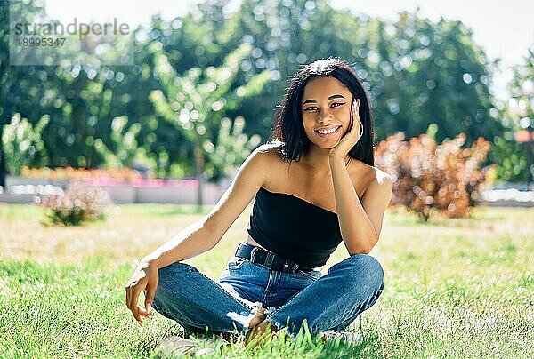 Hübsche lächelnde afroamerikanische Frau posiert im Park auf grünem Gras sitzend. Entspannen  Freizeit  weibliche Schönheit Konzept
