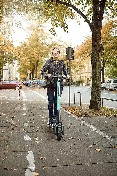 Junge glückliche blonde Frau fährt mit einem Elektroroller in der Stadt  lächelt in die Kamera  im Herbst