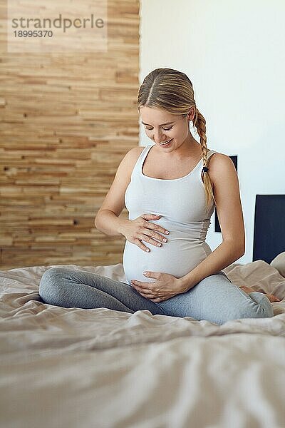 Glückliche junge schwangere Frau  die mit ihrem ungeborenen Kind auf einem Bett sitzt und ihren Babybauch mit ihren Händen wiegt und mit einem zärtlichen Lächeln der Liebe nach unten schaut