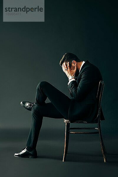 Trauriger  unglücklicher Mann im schwarzen Anzug  der auf einem Stuhl sitzt und das Gesicht mit zwei Händen verschließt. Profilansicht Porträt. Depression und Schmerz Konzept