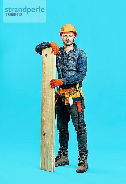 Ganzkörperporträt eines jungen  gut aussehenden Zimmermanns  der einen Stapel Holz vor einem blaün Hintergrund hält und zur Kamera schaut