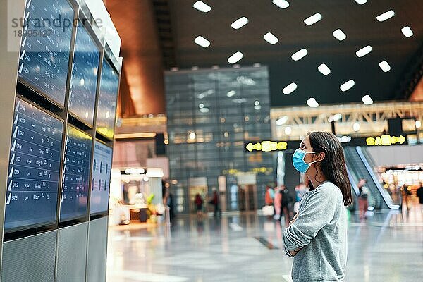 Frau mit Gesichtsmaske zum Schutz vor Viren schaut auf eine Informationstafel  um ihren Flug auf einem internationalen Flughafen zu überprüfen. Abflugtafel  Flugstatus