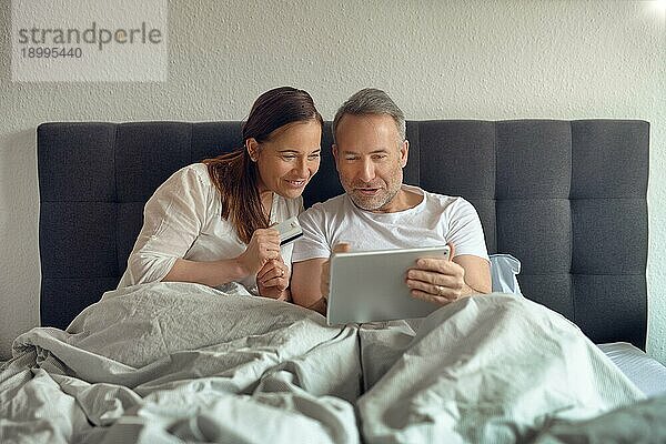 Glückliches Paar mittleren Alters  das morgens im Bett sitzt  Kaffee trinkt und mit einem digitalen Tablet online einkauft