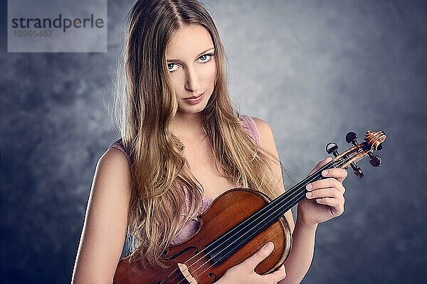 Attraktive junge Musikerin  die ihre Geige im Arm hält und lächelnd in die Kamera schaut  vor blauem Hintergrund