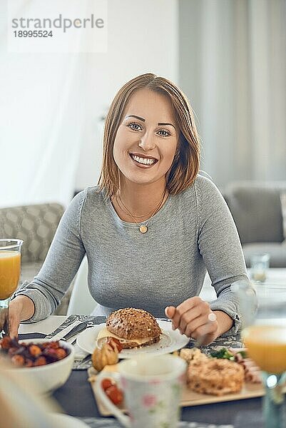 Glückliche attraktive Frau  die ein Sandwich zum Frühstück genießt  sitzt an einem gedeckten Tisch und lächelt in die Kamera
