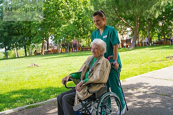 Eine ältere Frau mit der Krankenschwester auf einem Spaziergang durch den Garten eines Pflegeheims in einem Rollstuhl neben der Natur und den Bäumen