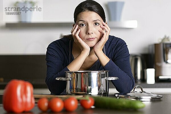 Unmotivierte attraktive junge Frau  die das Abendessen zubereitet und sich dabei auf das Kochfeld stützt  während sie mit lustlosem  mürrischem Gesichtsausdruck in ihrer Küche steht