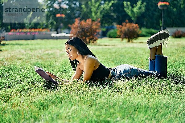 Schöne afroamerikanische Frau liest Buch im Gras liegend im Park. Entspannen und Hobby Konzept