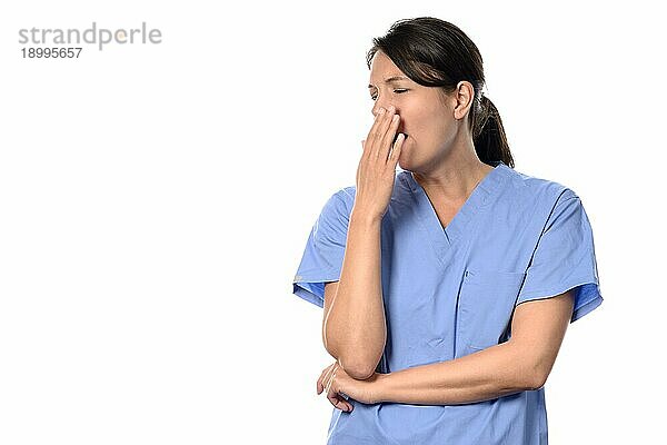 Erschöpfte attraktive Ärztin oder Krankenschwester in blauem Kittel  die nach einer langen  anstrengenden Schicht gähnend mit der Hand vor dem Mund steht  vor weißem Hintergrund