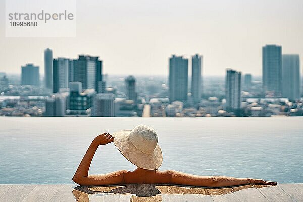 Rückenansicht der jungen Frau in Hut entspannend im Schwimmbad auf dem Dach des Hotels und genießen Stadtbild. Sommerurlaub  Luxuskonzept