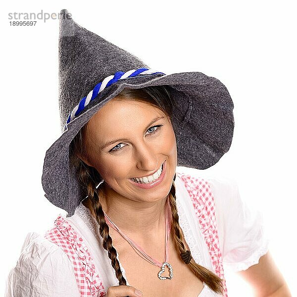 Schöne glückliche junge Frau mit geflochtenen Haaren  die ein traditionelles deutsches Dirndl und einen rustikalen Hut trägt und in die Kamera lächelt  vor weißem Hintergrund