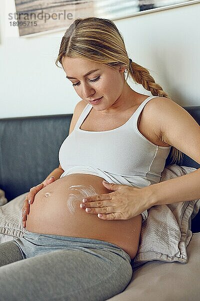 Junge schwangere Frau  die ihren Bauch mit Feuchtigkeitscreme einreibt  um ihre Haut mit Feuchtigkeit zu versorgen und die Gefahr von Dehnungsstreifen nach der Entbindung zu verringern