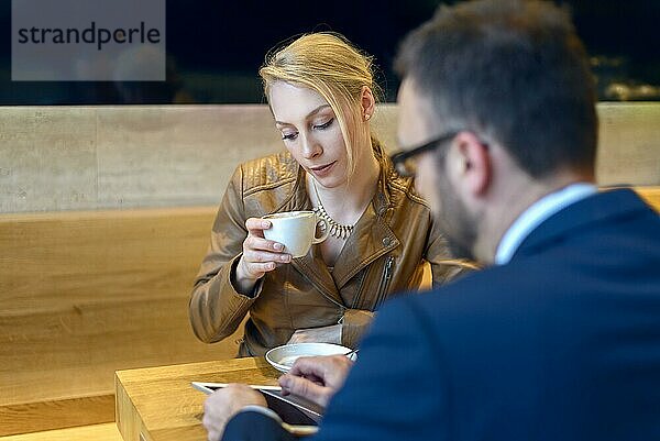 Geschäftsmann und Frau sitzen zusammen in einer Cafeteria und trinken Kaffee  wobei der Mann etwas auf seinem Tablet zeigt
