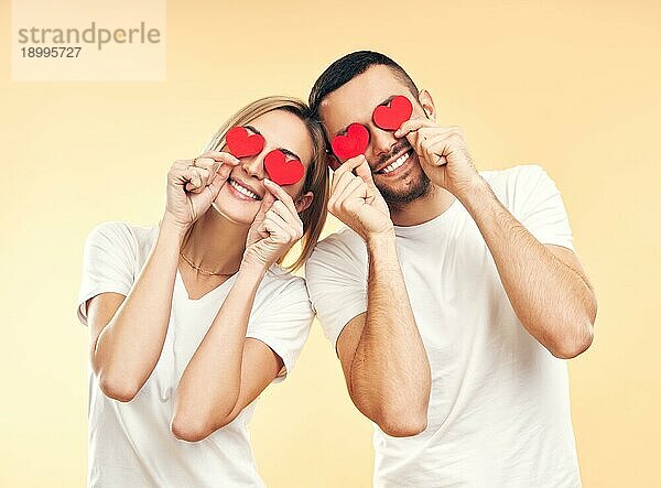 Happy couple have fun holding small paper hearts by their eyes over studio background. Liebe  Beziehung und romantisches Konzept