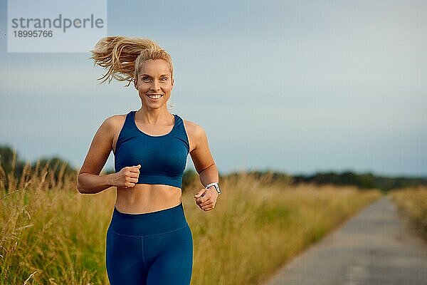 Fitte  gesunde junge Frau  die auf einer Landstraße joggt und mit einem glücklichen Lächeln voller Vitalität in einem aktiven Lebensstilkonzept an der Kamera vorbeigeht