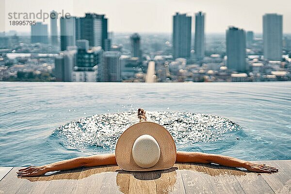 Rückansicht einer jungen Frau mit Hut  die sich im Schwimmbad auf dem Dach eines Hotels entspannt und das Stadtbild genießt. Sommerurlaub  Erholung Konzept