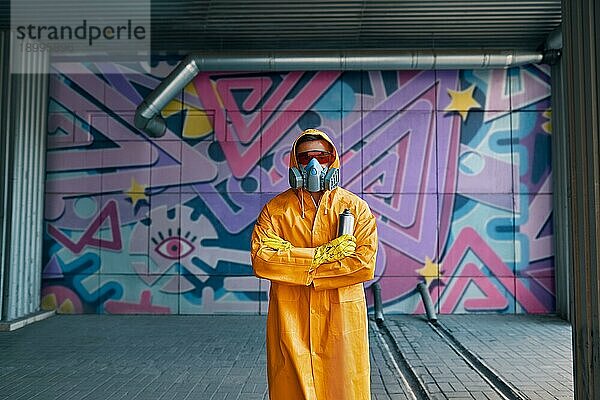 Graffitimaler mit Atemschutzmaske steht in der Nähe der Wand mit seinen Bildern und schaut in die Kamera. Street Art Konzept
