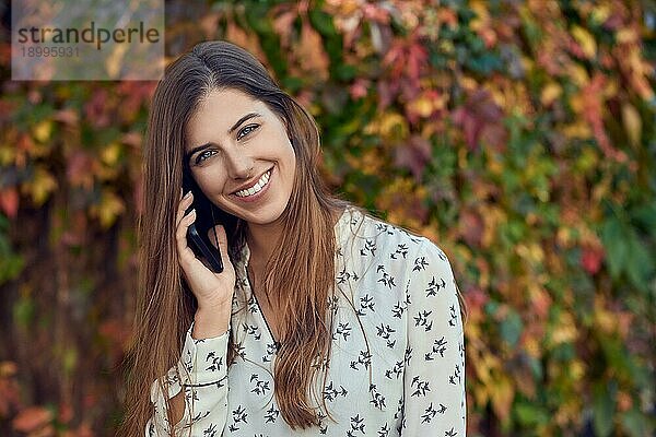 Lächelnde junge Frau  die im Herbst vor einer bunten Kletterpflanze steht und fröhlich in die Kamera lächelt  während sie mit dem Handy telefoniert