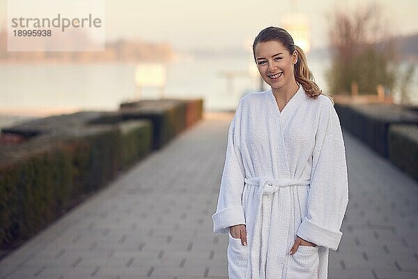 Lächelnde junge Frau im weißen Bademantel steht im Freien auf einem gepflasterten Weg  der zu einem ruhigen See führt  mit den Händen in den Taschen in einem Wellness und Spakonzept