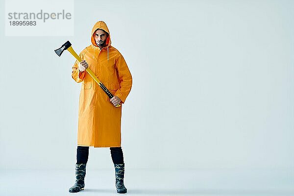 Ganzkörperporträt eines jungen  gut aussehenden Mannes in einem hellen Regenmantel  der eine große Axt hält  mit Kopierraum. Brutaler Männchen  Holzfäller  Fischer mit Axt