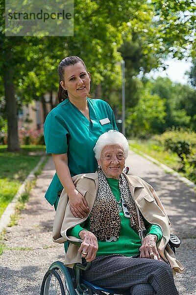 Eine ältere Frau mit der Krankenschwester bei einem Spaziergang im Garten eines Pflegeheims im Rollstuhl