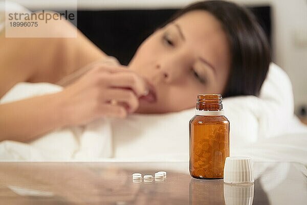 Eine Frau  die an einer Krankheit oder Schlaflosigkeit leidet  liegt im Bett und ist im Begriff  eine Tablette einzunehmen  während auf dem Nachttisch ein Fläschchen mit Medikamenten steht  wobei der Fokus auf dem Tablettenfläschchen liegt