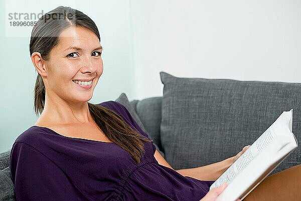 Lächelnde schöne junge Frau  die sich in ihrem Wohnzimmer auf einem Sofa entspannt und ein Buch liest  Nahaufnahme Porträt