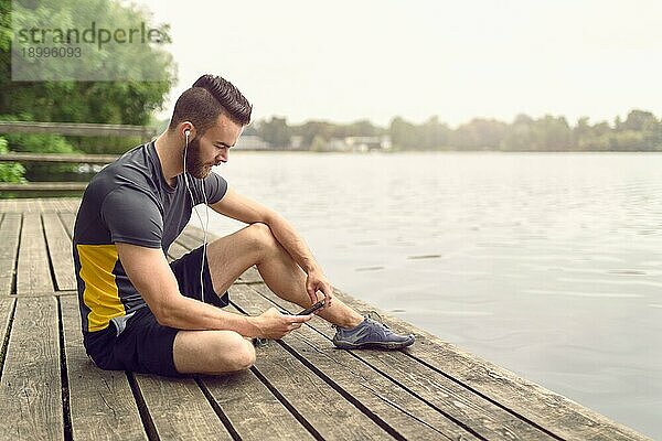 Attraktiver  bärtiger junger Mann  der sich auf einer Holzterrasse mit Blick auf einen ruhigen See entspannt und mit seinem Handy in der Hand lächelnd in die Kamera schaut