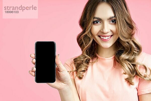 Close up Porträt der schönen lächelnden Frau hält leeren Bildschirm Handy über rosa Hintergrund
