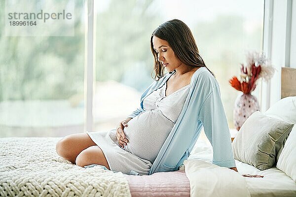 Porträt der jungen schwangeren Frau entspannen sittong im Bett und berühren ihren Bauch zu Hause. Mutterschaft  Menschen Konzept. Letzte Monate der Schwangerschaft