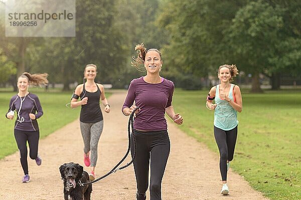 Halbkörperaufnahme einer sportlichen  hübschen jungen Frau  die in die Kamera lächelt  während sie mit anderen Mädchen im Park joggt  mit Kopierbereich auf der rechten Seite