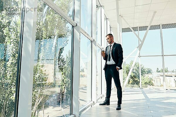 Full Länge Porträt der eleganten Geschäftsmann mit Tasse Kaffee in der Hand während der Pause in modernen kreativen Büro mit Kopie Raum. Entspannen  Erfolg Konzept