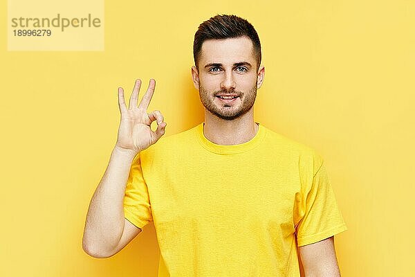 Lächelnd gut aussehend Mann tut ok Zeichen mit Hand und Finger über gelben Hintergrund. Erfolg Konzept