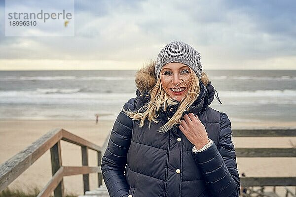 Frau mittleren Alters  die einem kalten Wintertag am Meer trotzt  steht auf einer Holzterrasse mit Blick auf den Strand an einem windigen Tag und lächelt glücklich in die Kamera