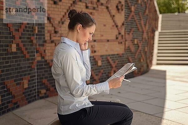 Frau  die in einem kommerziellen Foyer auf einer an der Wand befestigten Bank vor gemustertem Mauerwerk sitzt und mit ihrem Mobiltelefon spricht und ihre Notizen mit einem Tablet daneben bespricht  Konzept der Mobilität