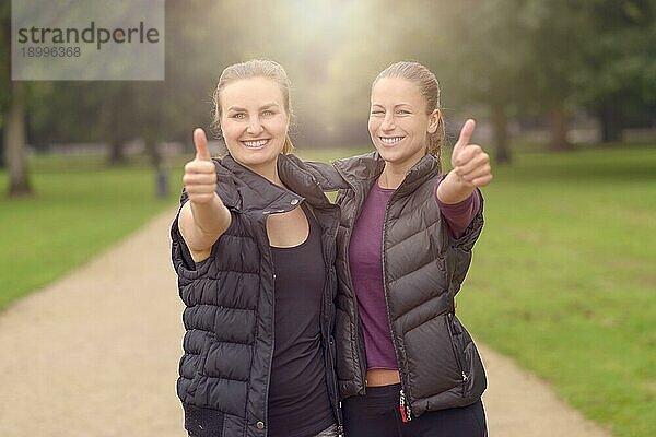 Halbkörperaufnahme von zwei glücklichen  gesunden Frauen im Park  die nach ihrem Training Daumen nach oben in die Kamera halten