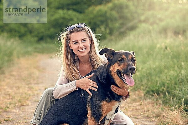 Glücklich lächelnder schwarzer Hund  der ein Laufgeschirr trägt und seiner hübschen jungen Besitzerin gegenübersitzt  die ihn mit einem liebevollen Lächeln im Freien auf dem Lande streichelt