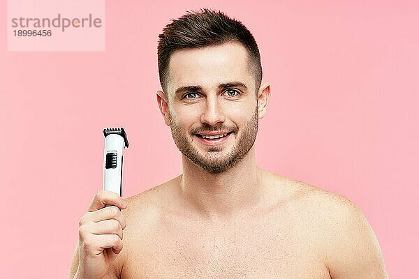 gutaussehender lächelnder Mann mit nacktem Oberkörper  der einen elektrischen Rasierapparat hält und in die Kamera auf rosa Hintergrund schaut