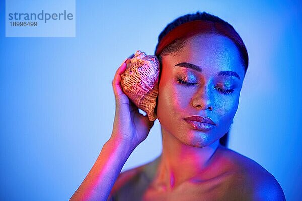 Hübsche afro Frau mit geschlossenen Augen drückte Muschel zu ihrem Ohr genießen und Träume mit Kopie Raum in Neonlicht. Sommerurlaub  Entspannung Konzept