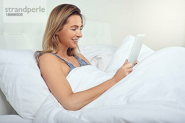 Attraktive Frau entspannt sich in einem bequemen neutralen weißen Bett mit einem Tabletcomputer in den Händen und einem freundlichen Lächeln