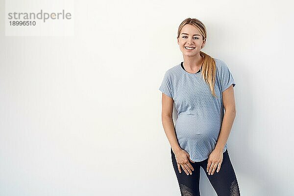 selbstbewusste  entspannte  attraktive  junge  schwangere Frau  die mit verschränkten Armen auf ihrem nackten Babybauch steht und mit einem freundlichen Lächeln in die Kamera blickt  auf weißem Grund mit Leerzeichen