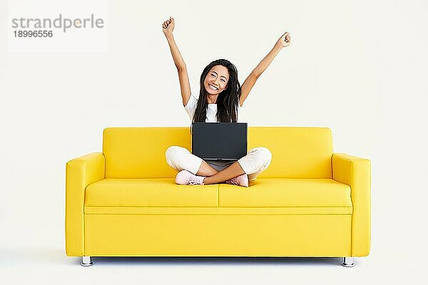 Glücklich lächelnde Frau mit erhobenen Armen sitzt auf gelber Couch mit Laptop und schaut in die Kamera auf weißem Hintergrund. Gewinnen  Feiern  Erfolgskonzept